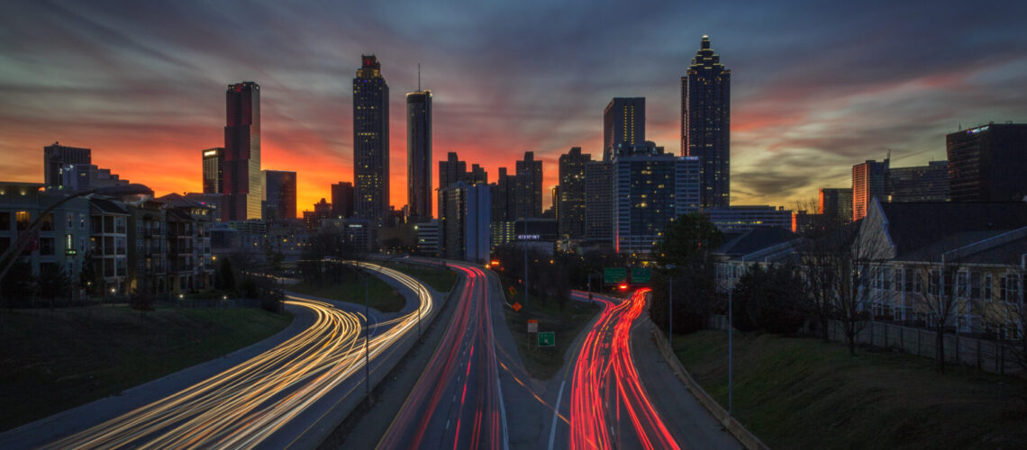 Long Exposure orange, pink, red, blue, purple sunset behind the Atlanta skyline from Jackson Street Bridge with streaked white and red car lights (company names have been edited out)